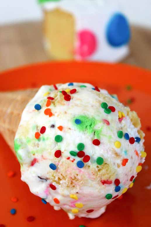 an ice cream cone filled with birthday cake ice cream lying sideways on an orange plate. 