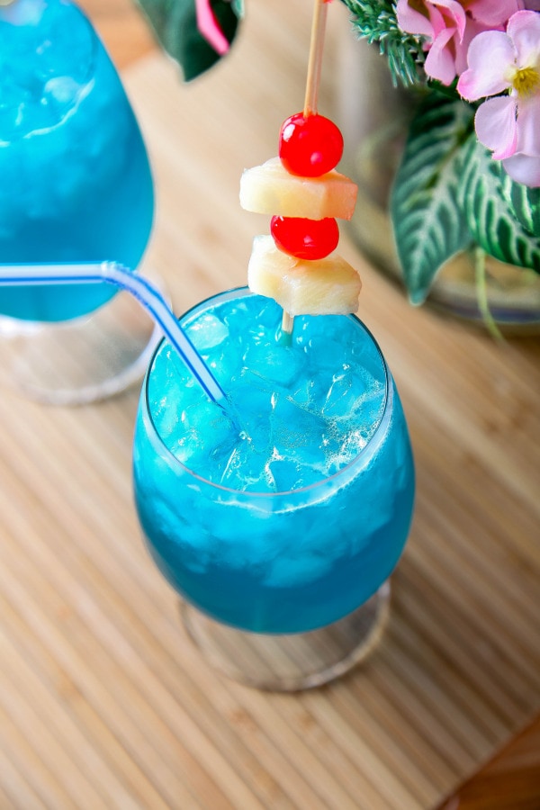 a closeup of a blue Hawaiian cocktail garnished with pineapple and maraschino cherries.