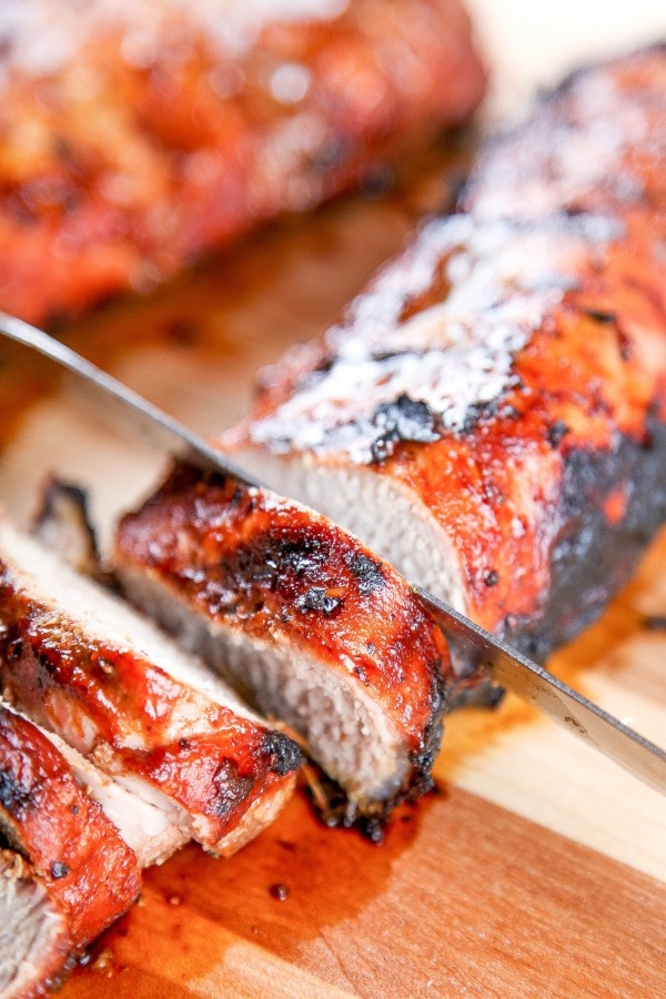 a knife slicing into a grilled BBQ pork tenderloin on a wooden cutting board.