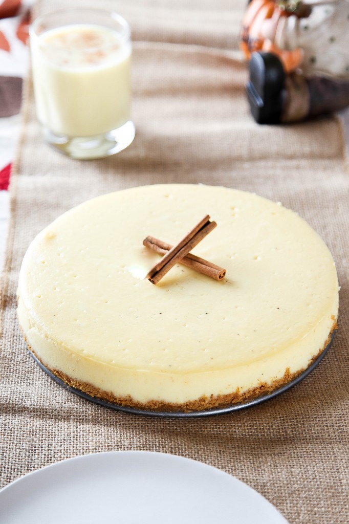 a whole eggnog cheesecake garnished 2 cinnamon sticks with a glass of eggnog in the background.