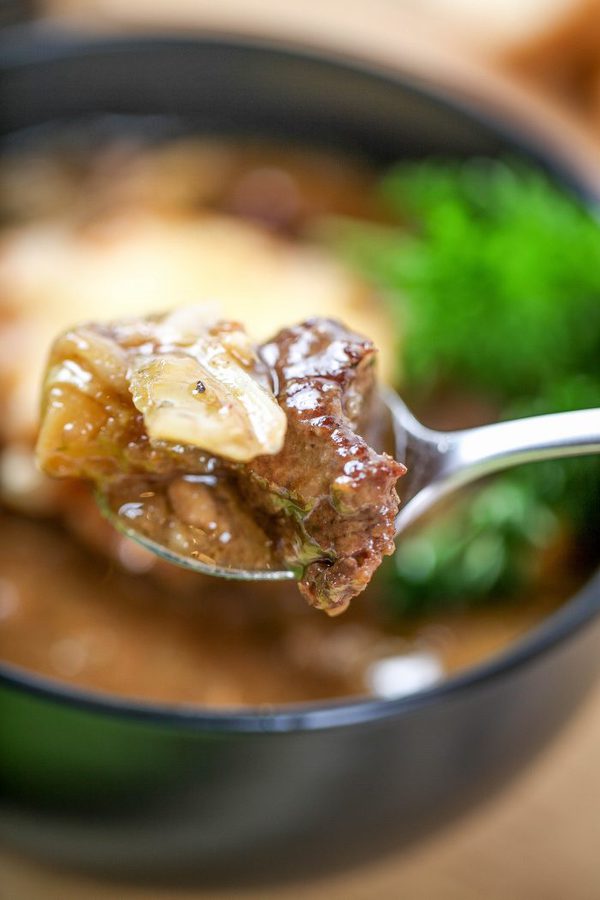 a spoon of beef stew containing beef, onion, and potato with the bowl in the background.