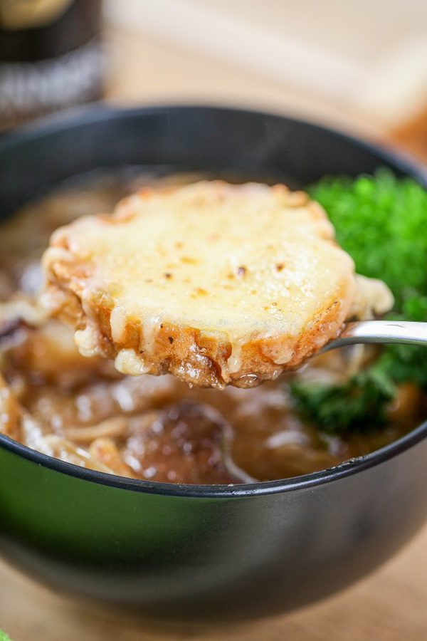 a spoon lifting a cheese covered baguette off the top of a bowl of Irish beef stew.