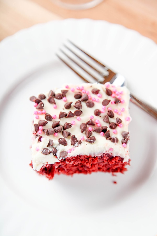 a slice of red velvet poke cake topped with cream cheese frosting, pink pearly sprinkles, and mini chocolate chips. 