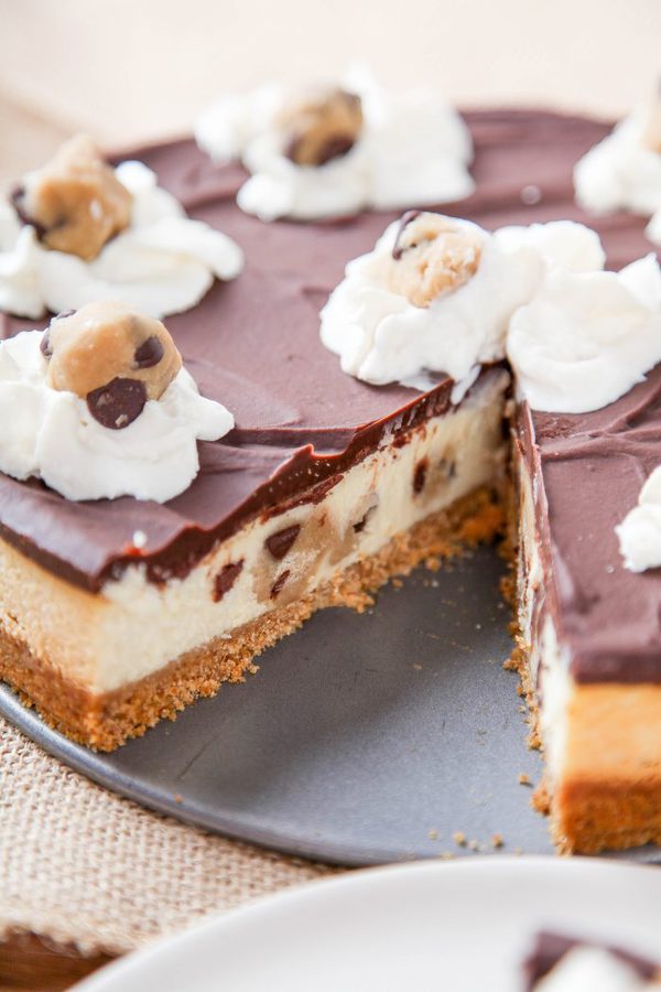 a whole cookie dough cheesecake topped with whipped cream and cookie dough with one slice cut out. 