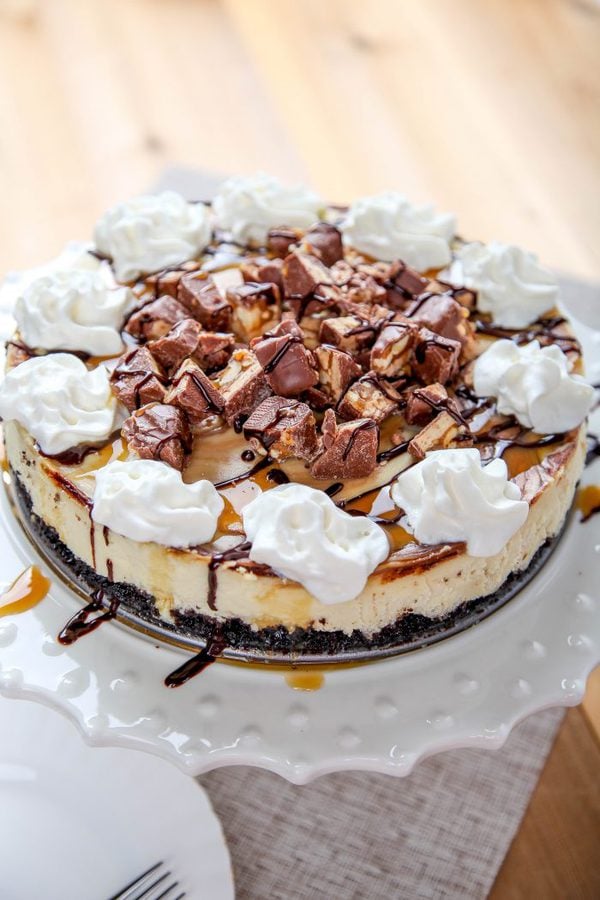 a whole snickers cheesecake topped with whipped cream, candy bars, caramel, and chocolate sauce. 