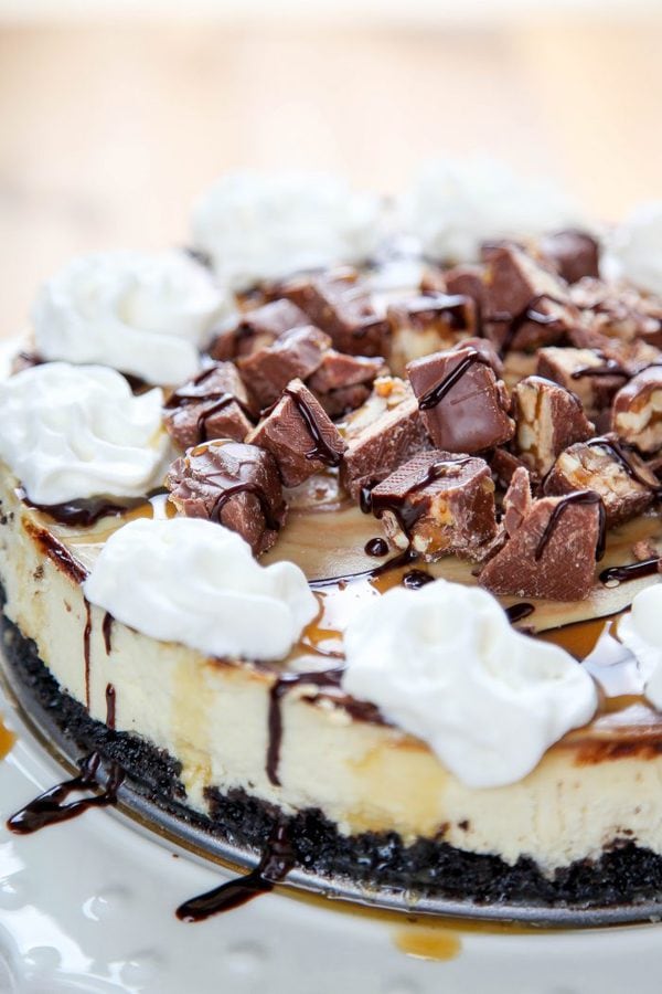 an oreo crust cheesecake topped with snickers, whipped cream, and caramel sauce.