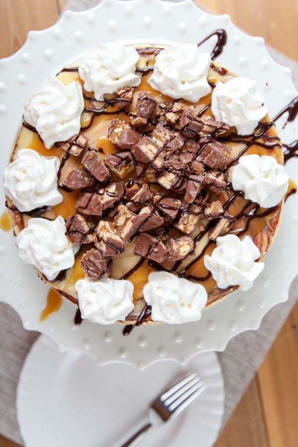 an overhead view of a snickers cheesecake with whipped cream on a white cake plate. 