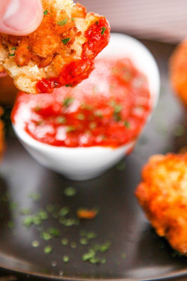 a deep fried macaroni and cheese fritter being dipped into a bowl of marinara sauce.