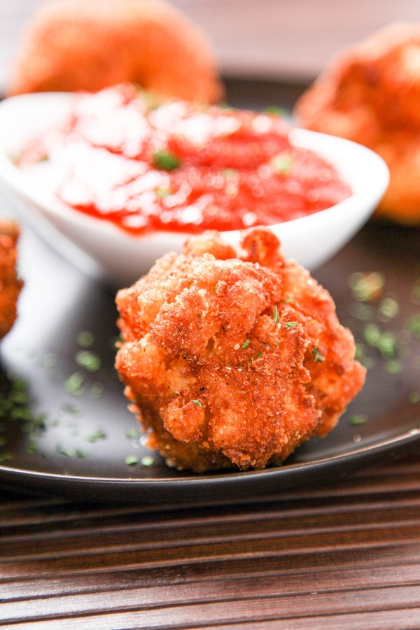 a black plate with a mac and cheese fritter with a bowl of marinara sauce in the background.