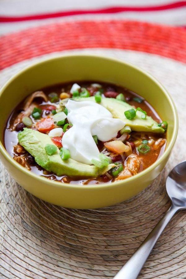 a bowl of taco soup topped with avocado and sour cream in a green bowl on a wicker placemat. 