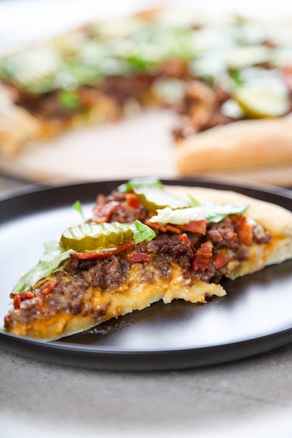 a side view of a slice of pizza topped with ground beef, bacon, cheese, pickles and lettuce.