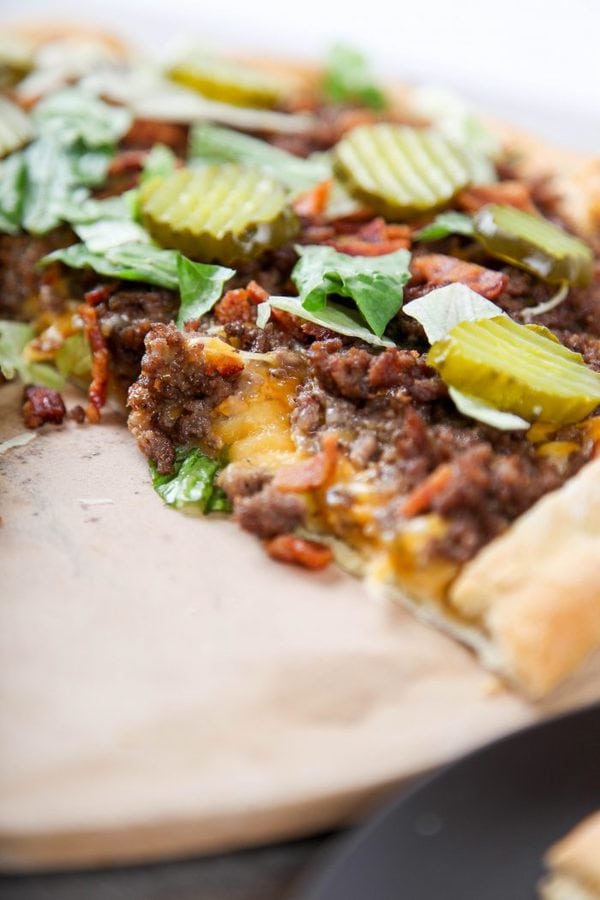a bacon cheeseburger pizza topped with pickles with one slice missing.