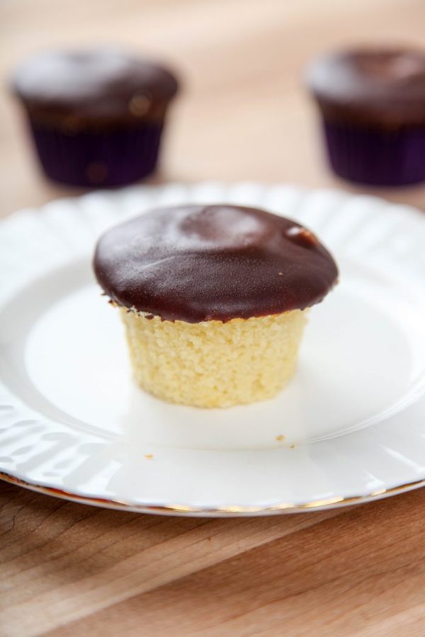 a boston cream pie cupcake on a white plate with gold edging with more cupcakes in the background.