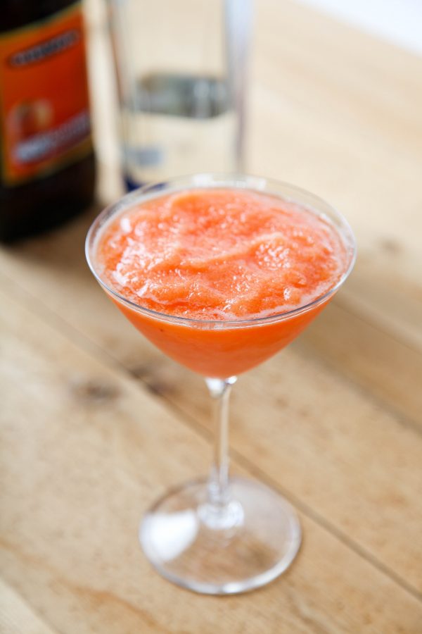 a peach bellini in a wine glass with peach schapps and sparkling wine bottles in the background.