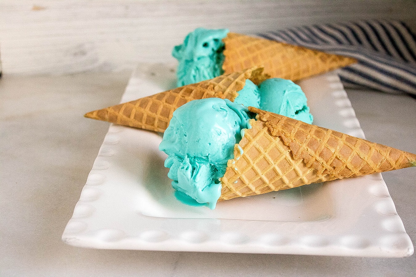 three waffle cons with blue ice cream on a white plate with a dish towel in the background.