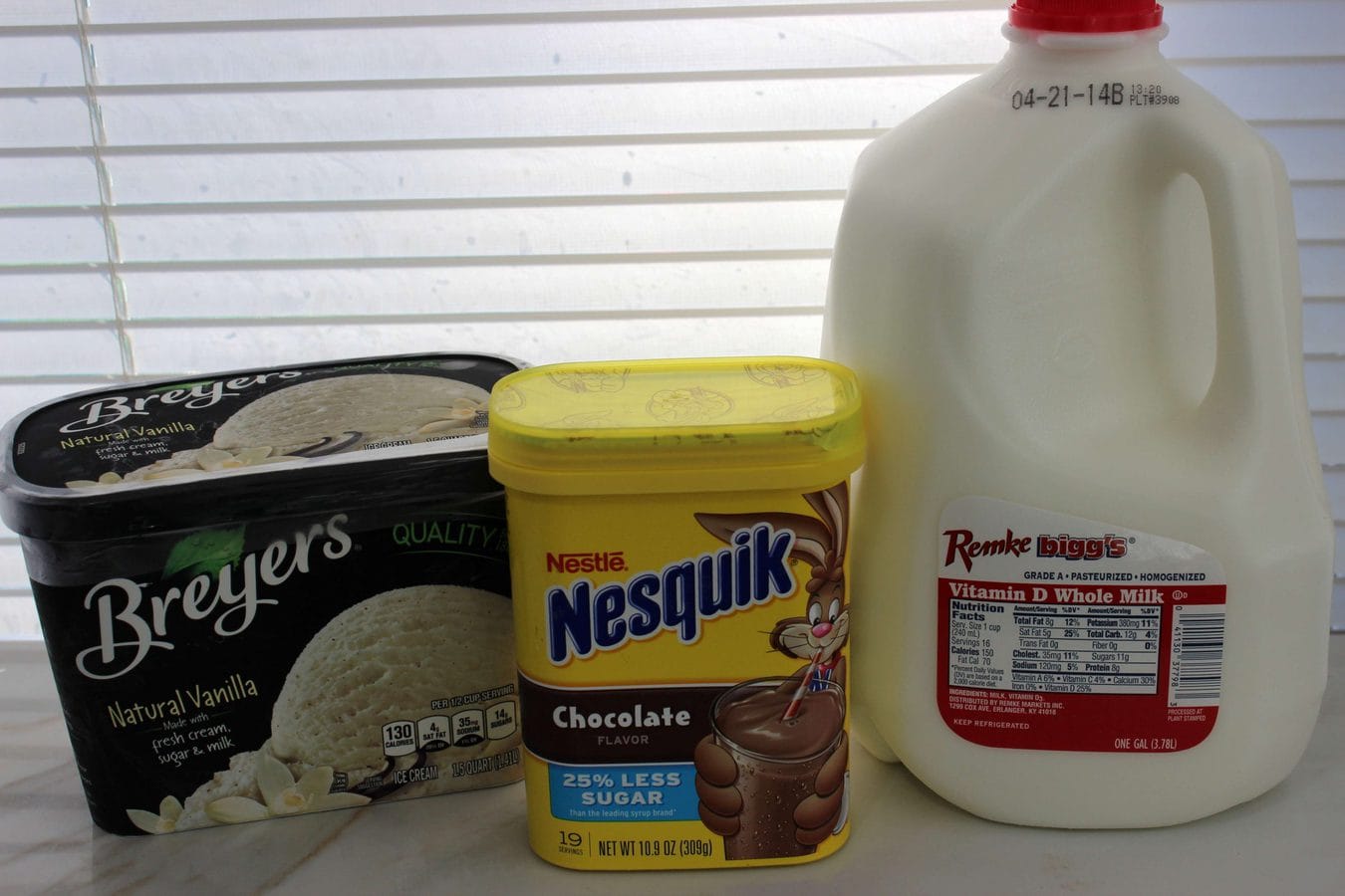 vanilla ice cream, nesquik powder, and a gallon of milk with blinds in the background. 