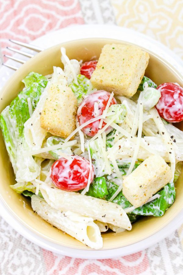 a yellow bowl of caesar pasta salad with penne noodles, cherry tomatoes, and croutons. 
