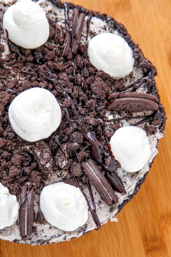 an overhead shot of a whole oreo cheesecake topped with oreo cookie crumbs, whole oreos, and dollops of whipped cream.