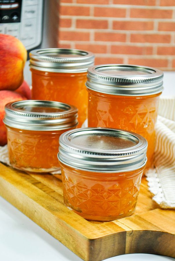 2 tall and 2 short glass mason jars filled with peach jam with fresh peaches in the background.