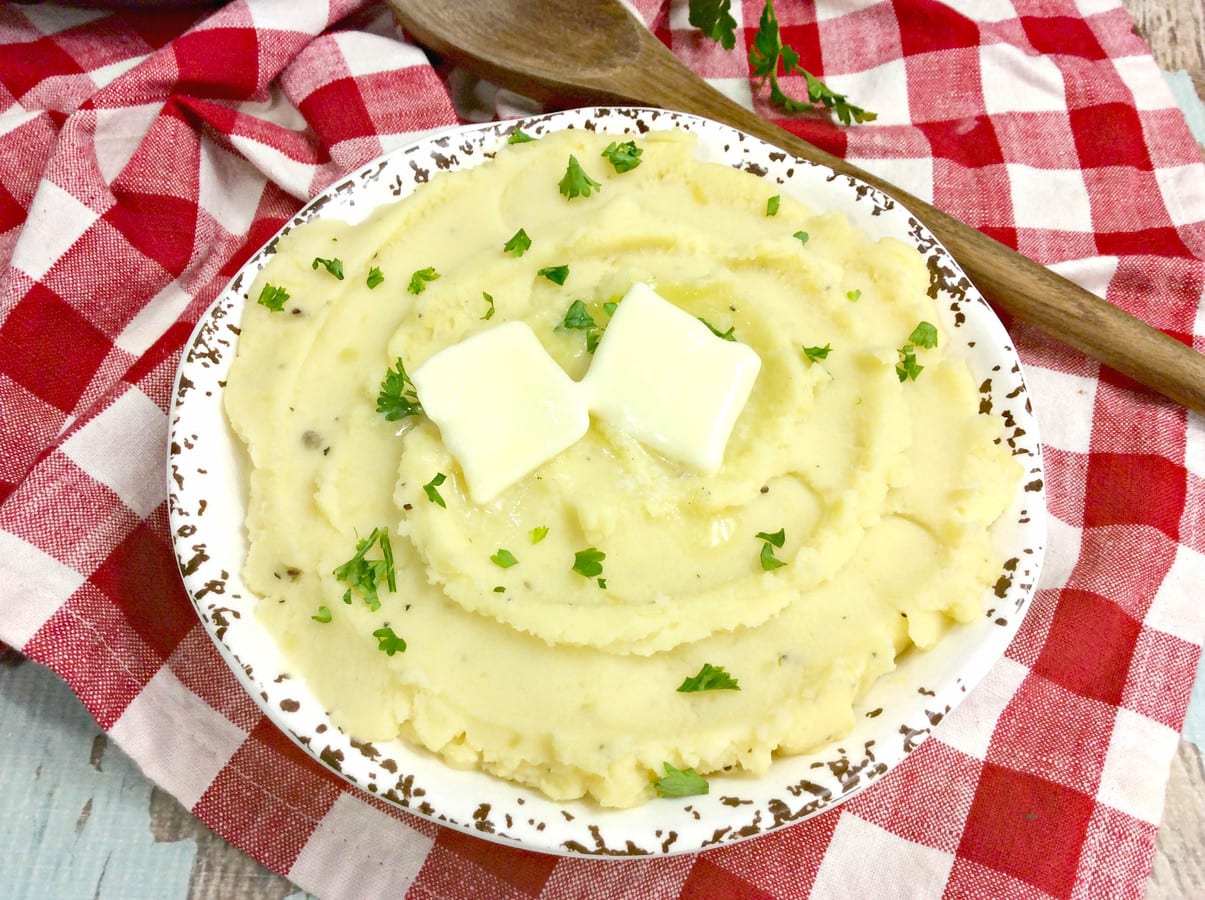 A rustic, white ceramic bowl filled with creamy mashed potatoes, topped with two pats of butter and sprinkled with chopped parsley, sits on a red and white checkered cloth. A wooden spoon lies beside the bowl.
