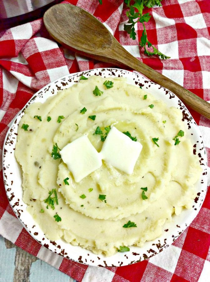 a bowl of mashed potatoes topped with 2 pats of butter on a checkered tablecloth.