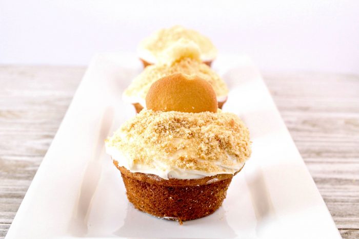 three banana pudding cupcakes on a white plate topped with frosting and vanilla wafers. 