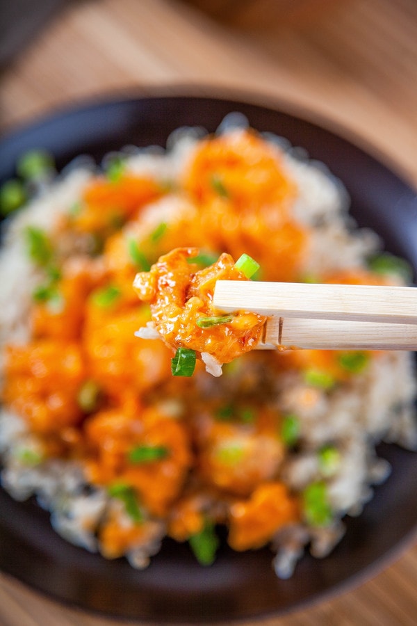 a close up of chopsticks holding one bang bang shrimp with a bowl of shrimp in the background.
