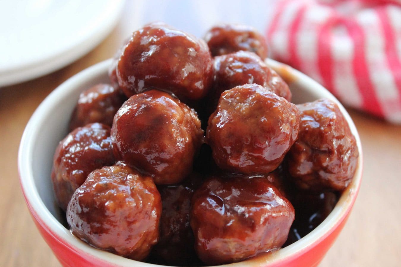 a closeup of meatballs in a chili and grape jelly sauce with a red tea towel in the background.