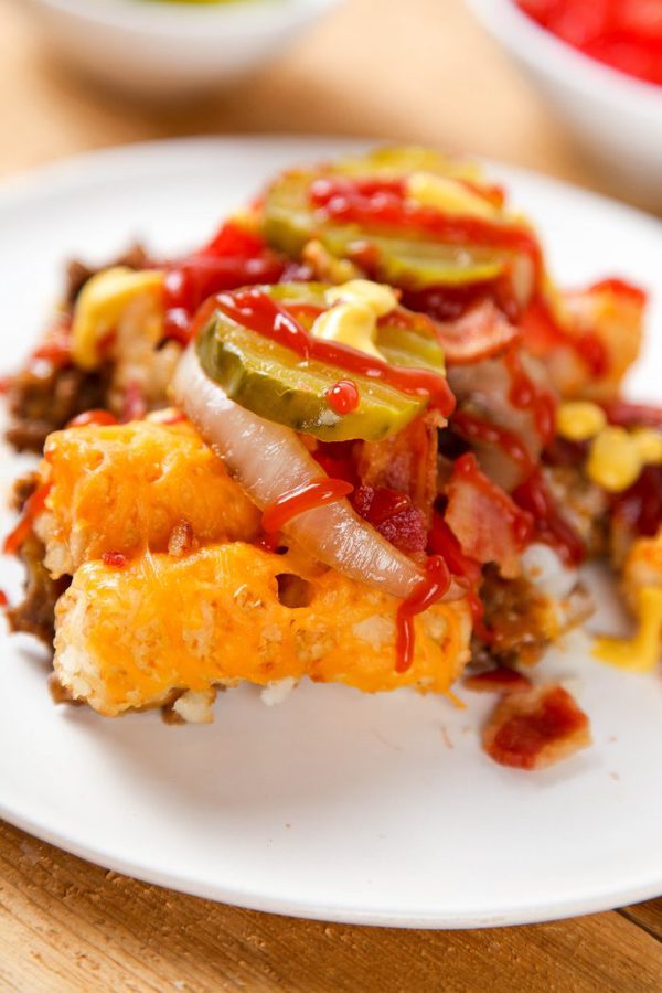 a close up of a cheeseburger casserole topped with cheesy tater tots and pickles. 