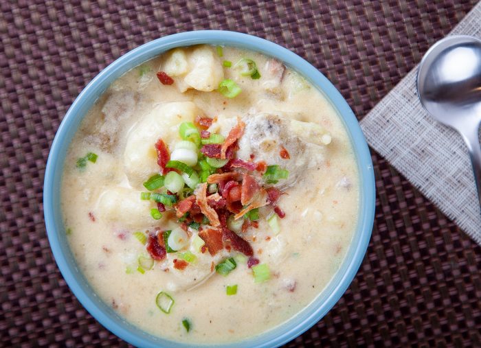 an overhead bowl of baked potato soup topped with bacon and green onions with a spoon to the right.