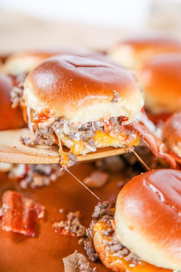 a wooden spatula lifting out slider rolls with hamburger meat, cheese, and bacon.