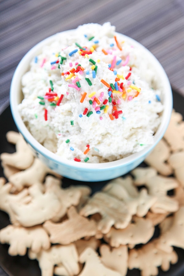 A bowl of cake batter dip topped with rainbow sprinkles with lots of animal crackers in front. 