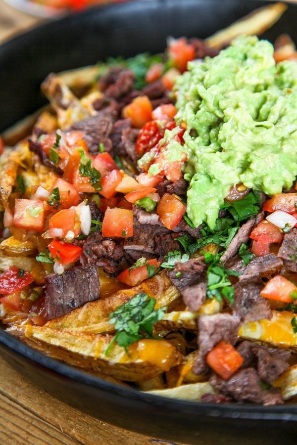 a closeup image of sliced carne asada steak, pico de gallo, and guacamole on top of homemade french fries.