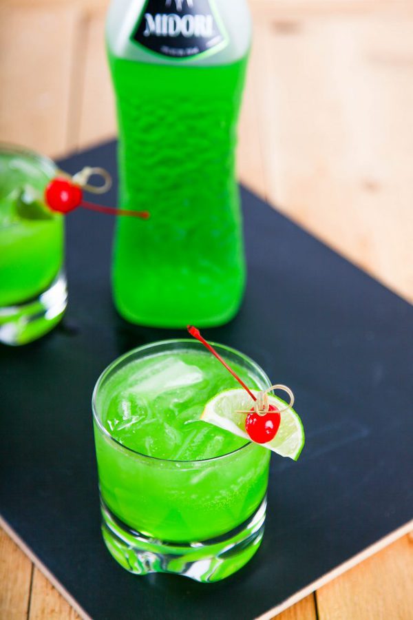 a overhead view of a midori drink with lime and a cherry with a bottle of midori in the background.
