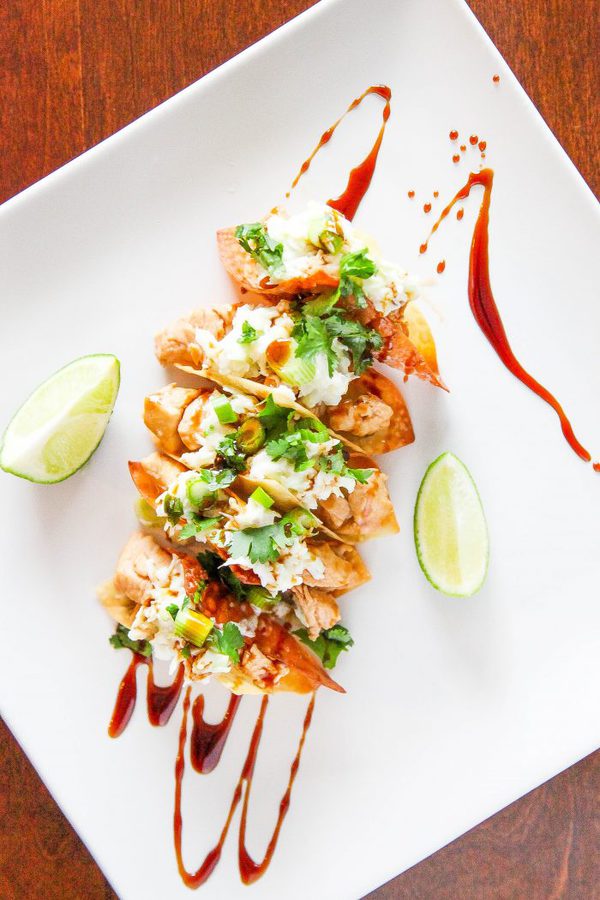 an overhead view of a plate of 5 chicken wonton tacos with a soy sauce drizzle and lime slices.