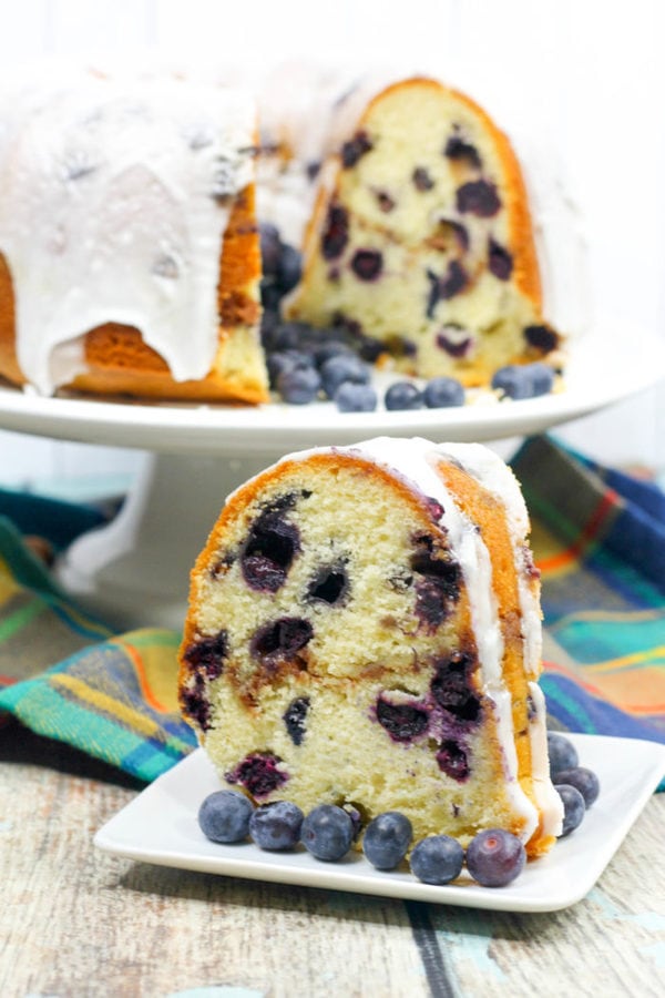 a large slice of blueberry bundt cake surrounded by fresh blueberries with the full cake in the background. 