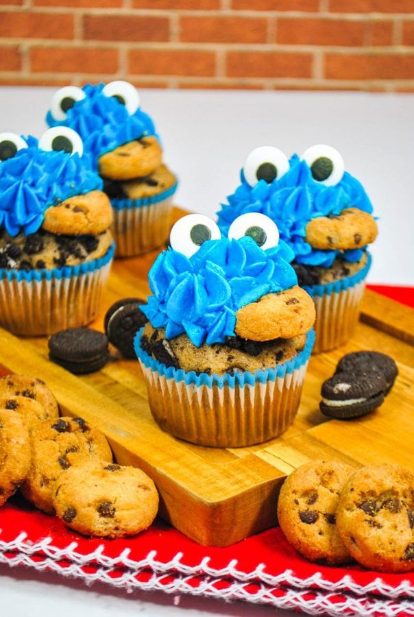 4 cookie  monster cupcakes eating with candy eyes and a chocolate chip cookie in their mouths.