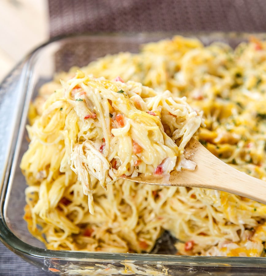 A wooden spoon lifting chicken spaghetti out of a glass casserole dish.