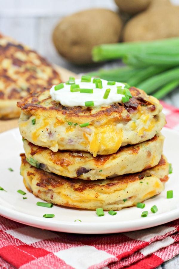 three mashed potato cakes on a white plate with green onions and potatoes in the background.