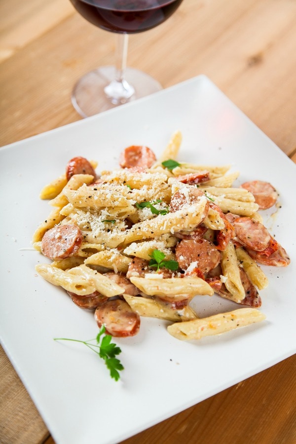 penne pasta with sausage and sun dried tomatoes on a white place with a glass of wine.
