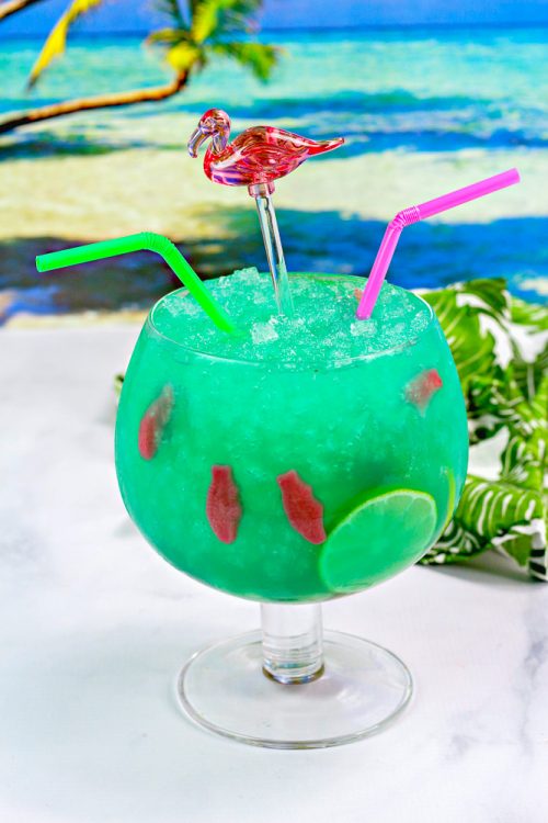 a fish bowl cocktail glass filled with a green mixed drink and swedish fish.