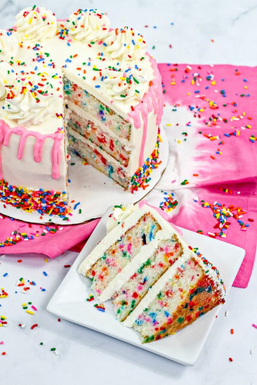 an overhead shot of a funfetti cake with one slice taken out of it, which sits nearby on a white plate. 
