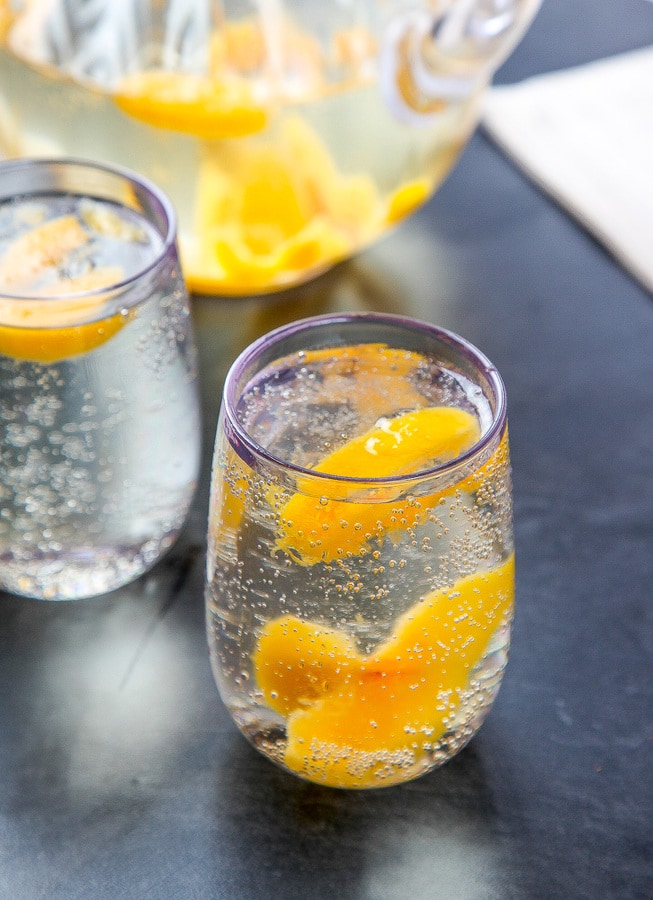 a glass of white wine sangria with peaches in a clear glass with a pitcher in the background.