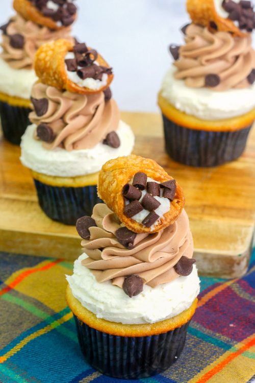 4 cannoli cupcakes on a wooden cutting board and a multi colored dish towel