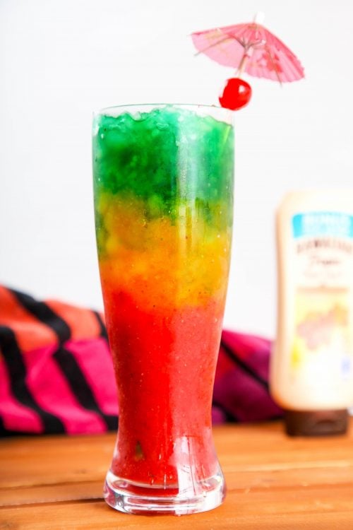 a tall cocktail glass filled with red, yellow, and green layers garnished with a cherry.