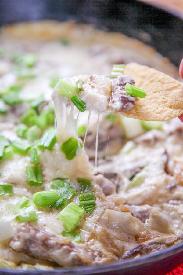 A torilla chip dipping into philly cheesesteak dip in a cast iron skillet.
