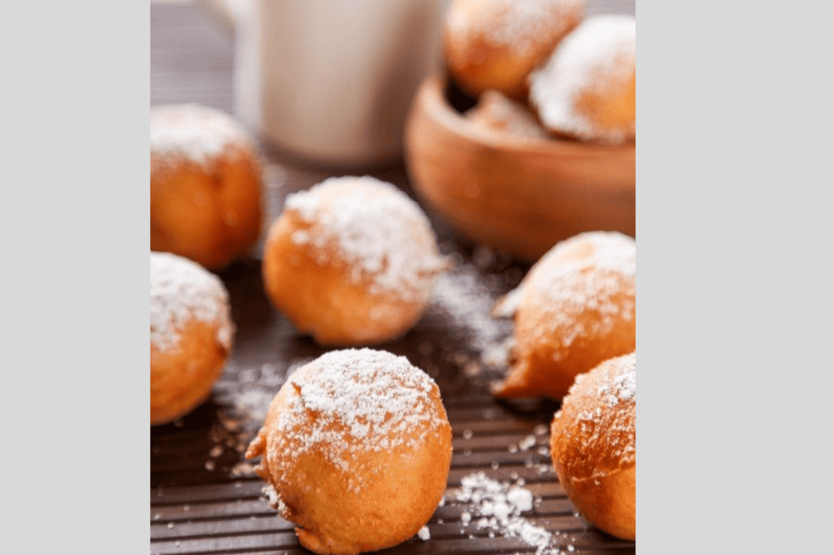 Fried Italian donut holes topped with powdered sugar with a coffee pitcher in the background.