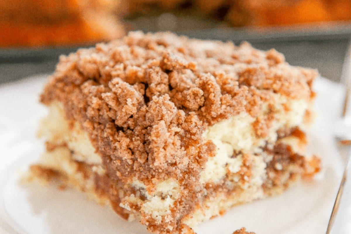 A close-up of a slice of coffee cake with a crumbly streusel topping. The cake appears moist with a light, fluffy texture and is shown on a white plate in a well-lit setting.