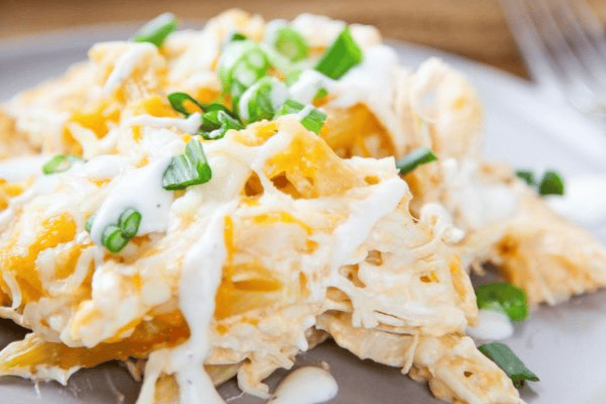 A close-up of a creamy shredded chicken dish topped with melted cheese, chopped green onions, and drizzled with white sauce. The dish appears to be served on a grey plate with a blurred background.