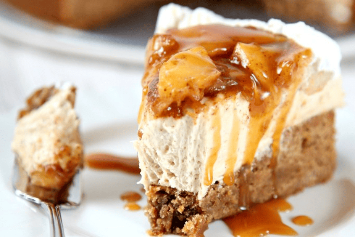 A close-up of a slice of cheesecake topped with caramel sauce and chunks of fruit. The cheesecake rests on a crumbly crust, and a spoon lies beside it with a bite of the dessert on it. The background is slightly blurred, focusing attention on the cheesecake.